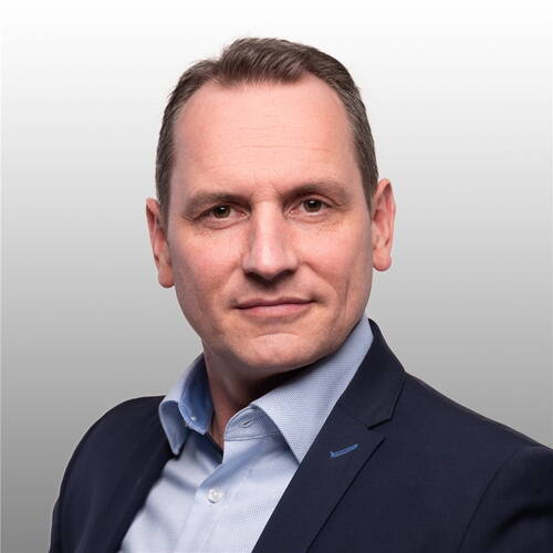Mittelalterlicher Mann im Business-Anzug mit hellblauem Hemd blickt ernst und direkt in die Kamera vor neutral grauem Hintergrund
