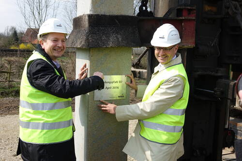 Zwei Bauingenieure in Sicherheitswesten und Helmen befestigen lächelnd eine Hinweistafel an einem frisch gesetzten Betonpfahl auf einer Baustelle im Freien.