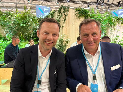 Zwei lächelnde Männer im Anzug mit Konferenz-Badges sitzen in einer begrünten Messe-Lounge, während im Hintergrund weitere Teilnehmende in entspannter Atmosphäre miteinander sprechen.