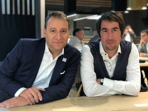 Zwei Männer in Businesskleidung sitzen entspannt nebeneinander an einem Konferenztisch und blicken lächelnd in die Kamera, während im Hintergrund weitere Teilnehmende in Gespräche vertieft sind.
