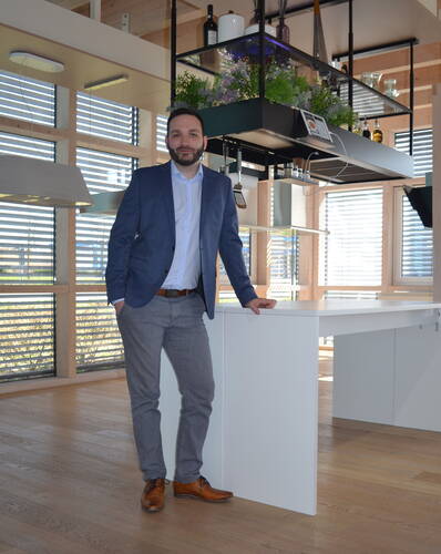 Geschäftsmann im blauen Sakko lehnt entspannt an einer modernen weißen Kücheninsel in einem hellen, holzbetonten Büro mit hängendem Regal und Pflanzen über der Arbeitsfläche