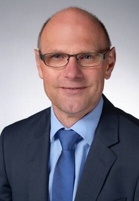 Glatzköpfiger Mann mit Brille im dunkelblauen Anzug und blauer Krawatte blickt freundlich in die Kamera vor neutralem Hintergrund