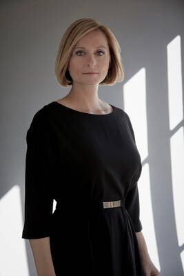 Frau mit blondem Bob und schwarzem Kleid steht ernst blickend im weichen Seitenlicht vor einer grauen Wand mit hellen Fensterschatten