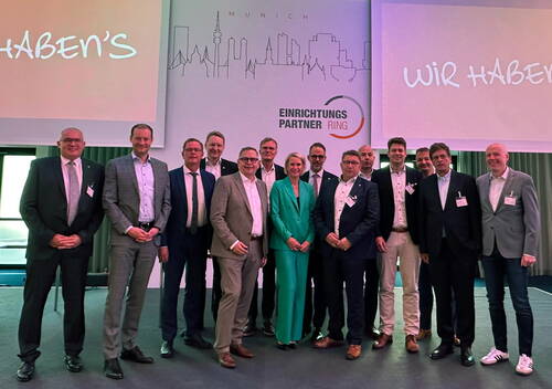 Gruppe von Geschäftsleuten in Anzügen und Namensschildern posiert auf einer Bühnenfläche vor einer Leinwand mit dem Schriftzug „Einrichtungspartner Ring“ und einer stilisierten Skyline von München.