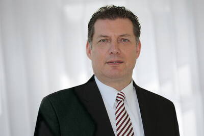 Mittelalterlicher Mann im dunklen Anzug mit weißem Hemd und rot-weiß gestreifter Krawatte steht vor einem hellen, unscharfen Hintergrund und blickt ernst in die Kamera.