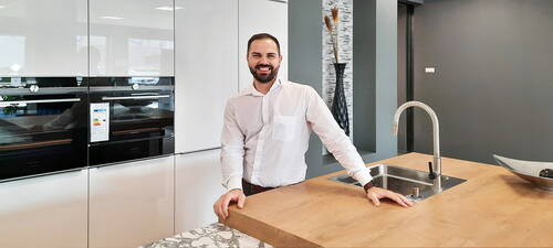 Lächelnder Mann im weißen Hemd lehnt sich auf eine moderne Kücheninsel mit Edelstahlspüle und steht vor eingebauten schwarzen Backöfen in einer hellen Designküche.