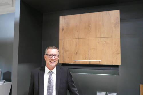 Lächelnder Mann im Anzug und Krawatte steht vor einem modernen Küchenoberschrank aus hellem Holz vor dunkler Wand.