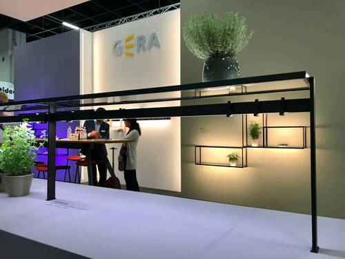 Modern gestalteter Messestand mit beleuchteten Regalsystemen, Pflanzen in Töpfen und zwei Personen, die an einem Stehtisch unter einer schwarzen, rahmenartigen Lichtkonstruktion sprechen.