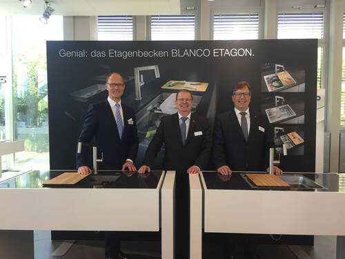 Drei Anzugträger präsentieren in einer modernen Ausstellungshalle das Etagenbecken BLANCO ETAGON an zwei Kücheninseln vor einer großformatigen Produktwand.
