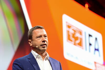Redner im blauen Anzug mit Headset-Mikrofon steht vor einer großen orangefarbenen IFA-Bühnenleinwand