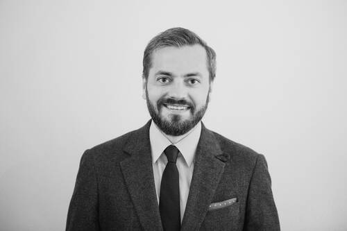 Lächelnder Mann im Anzug mit Krawatte und Einstecktuch steht vor einem hellen, neutralen Hintergrund.