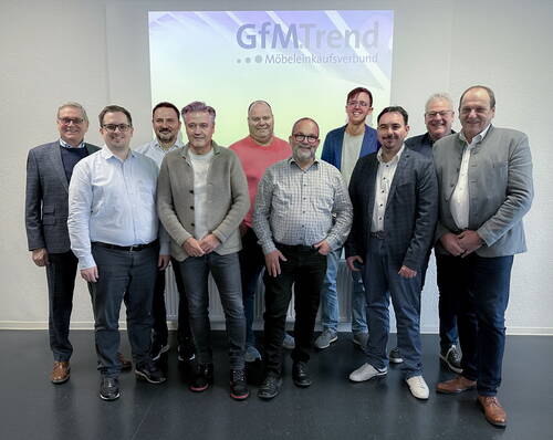 Gruppe von neun Männern in Business-Kleidung steht lächelnd vor einer Leinwand mit dem Logo „GfMTrend – Möbeleinkaufsverbund“ in einem Tagungsraum