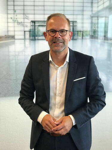 Mittelalterlicher Mann im dunklen Anzug mit weißem Hemd und Brille steht lächelnd in einer modernen, lichtdurchfluteten Empfangshalle mit Glasfront.