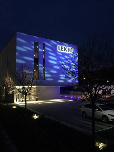 Modernes Firmengebäude mit der Leuchtreklame „LEICHT“, dessen Fassade nachts von blauen, kreisförmigen Lichtmustern und einem beleuchteten Parkplatz mit geparktem Auto angestrahlt wird.