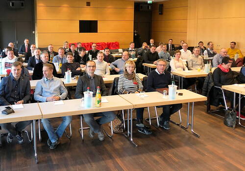 Teilnehmende einer geschäftlichen Schulung sitzen in Reihen an Tischen in einem Konferenzraum und blicken aufmerksam nach vorn.