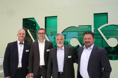 Vier geschäftlich gekleidete Männer posieren lächelnd nebeneinander vor einer grün beleuchteten Glaswand mit großem stilisiertem Logo.