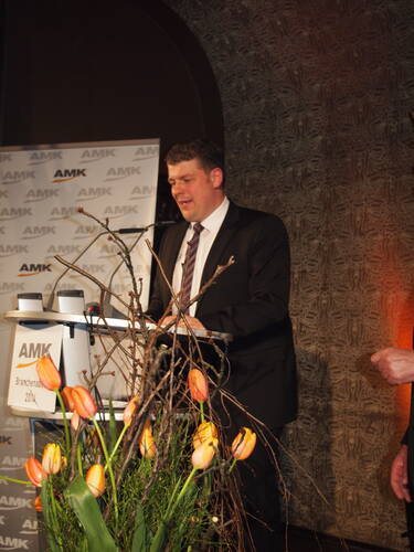Mann im Anzug hält eine Rede am AMK-Rednerpult, im Vordergrund ein großer Strauß orangefarbener Tulpen und Zweige.