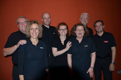 Sieben lächelnde Teammitglieder in schwarzen Poloshirts posieren vor einer orangefarbenen Wand und stehen dicht beieinander.