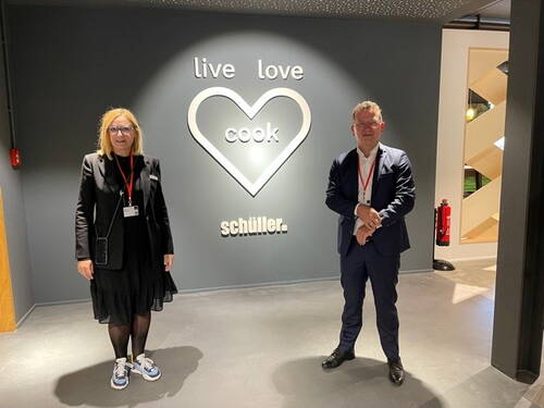 Zwei Geschäftsleute mit Besucherausweisen stehen in einer modernen Firmenlobby vor einer dunkelgrauen Wand mit großem Herzsymbol und dem Schriftzug „live love cook schüller.“