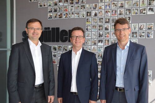Drei lächelnde Männer in Anzügen stehen nebeneinander vor einer Wand mit zahlreichen Polaroid-Fotos von Mitarbeitenden in einem modernen Büro.
