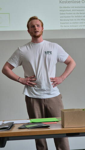 Junger Mann mit LIFE‑Affirming‑Activism‑T-Shirt hält einen Vortrag vor einer Leinwand mit Text über kostenlose Onlineberatung und steht mit Händen in der Hüfte hinter einem Tisch mit Unterlagen und Karton.