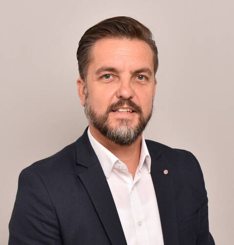 Mittelalterlicher Mann mit kurzem, gegeltem Haar und Vollbart trägt dunklen Anzug und weißes Hemd und schaut lächelnd in die Kamera vor neutralem Hintergrund