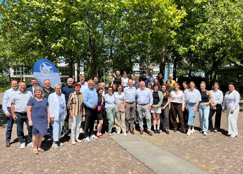 Große gemischte Gruppe aus jüngeren und älteren Personen posiert lächelnd bei sonnigem Wetter unter Bäumen auf einem gepflasterten Platz vor einem Verwaltungsgebäude mit blauem Veranstaltungsbanner.
