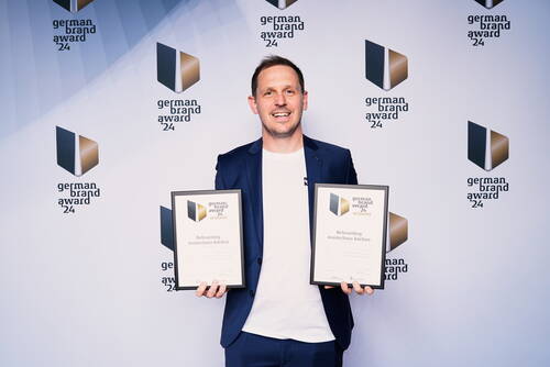Strahlender Preisträger im blauen Anzug hält bei der German Brand Award 2024 Verleihung zwei Urkunden für das Rebranding von Musterhaus Küchen vor Fotowand mit Award-Logos.