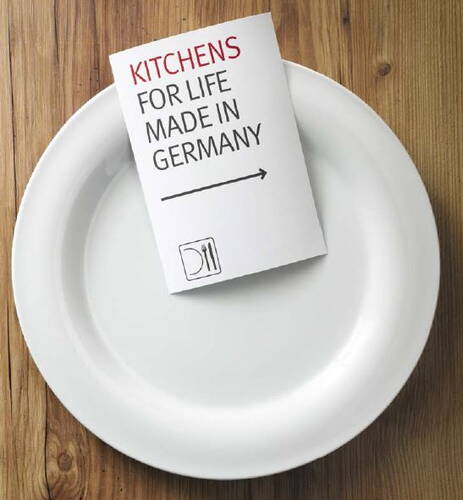 Ein weißer Teller auf Holztisch, darauf ein Flyer mit der Aufschrift „KITCHENS FOR LIFE MADE IN GERMANY“ und einem Pfeil nach rechts