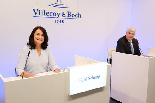 Zwei Führungskräfte sitzen an weißen Rednerpulten mit Mikrofonen in einem modernen Konferenzraum von Villeroy & Boch, während die namentlich gekennzeichnete Sprecherin Gabi Schupp lächelnd in die Kamera blickt.