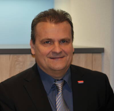 Mittelalterlicher Mann im Business-Anzug mit blauer Krawatte lächelt leicht in einem modernen Büroambiente.