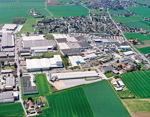 Luftaufnahme eines weitläufigen Industriegebiets am Stadtrand, umgeben von grünen landwirtschaftlichen Feldern und einem angrenzenden Wohnviertel.