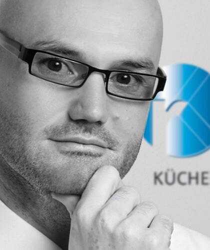 Nachdenklicher, glatzköpfiger Mann mit Brille im Porträt, der das Kinn stützend in die Kamera blickt, vor einem unscharfen blauen Firmenlogo im Hintergrund
