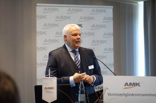 Geschäftsmann im Anzug hält eine Rede am Rednerpult während einer AMK‑Vereinsmitgliederversammlung in einem Konferenzraum.