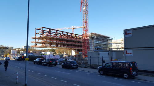 Große Baustelle eines mehrstöckigen Stahl- und Glasgebäudes mit rotem Kran neben befahrener Straße mit parkenden Autos bei klarem blauem Himmel
