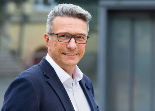 Reifer Geschäftsmann mit grauem Haar und Brille lächelt im Anzug vor einem modernen Gebäude in die Kamera