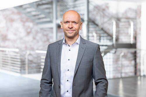 Glatzköpfiger Mann mittleren Alters im grauen Business-Anzug lächelt selbstbewusst in einer modernen Bürohalle mit Treppenaufgang im Hintergrund