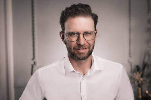 Mittelalterlicher Mann mit Brille und Bart lächelt freundlich in die Kamera und trägt ein weißes Hemd vor unscharfem, hellgrauem Hintergrund