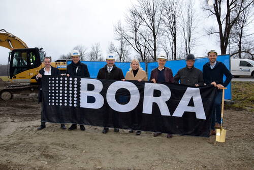 Gruppe von sieben Personen mit Bauhelmen hält ein großes schwarzes BORA-Banner vor einer Baustelle mit Bagger und blauem Bauzaun im Hintergrund
