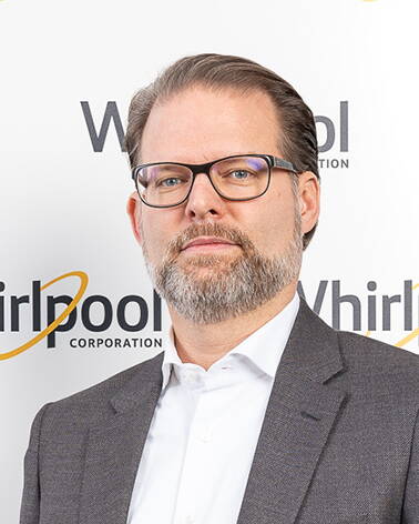 Mittelalter Mann mit Brille und grauem Vollbart im Anzug steht vor einer weißen Wand mit wiederholtem Whirlpool-Logo
