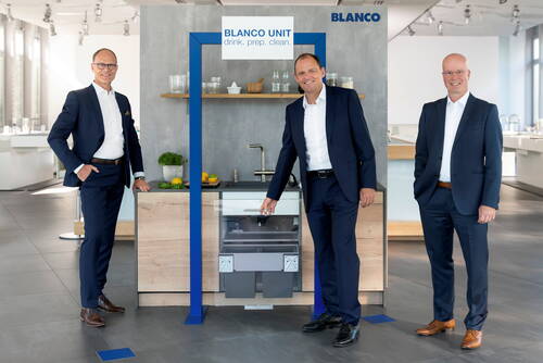 Drei Geschäftsleute in Anzügen präsentieren in einem modernen Showroom eine BLANCO Küchenzeile mit Spüle, Armatur und offenem Abfallsystem unter dem Schriftzug „BLANCO UNIT – drink. prep. clean.“