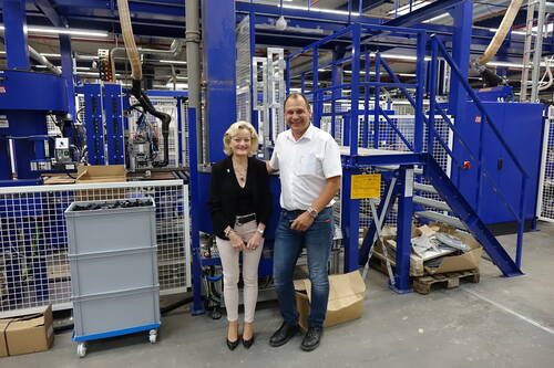 Zwei lächelnde Personen in Businesskleidung stehen in einer modernen Fabrikhalle vor blauen Industrieanlagen und Fördertechnik
