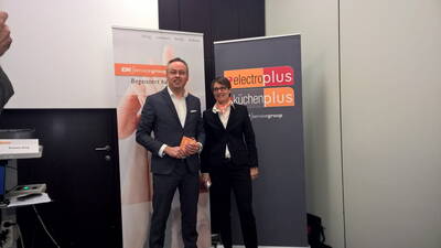 Zwei Geschäftsleute stehen lächelnd im Anzug zwischen EK/servicegroup- und electroplus küchenplus‑Rollups in einem Konferenzraum.
