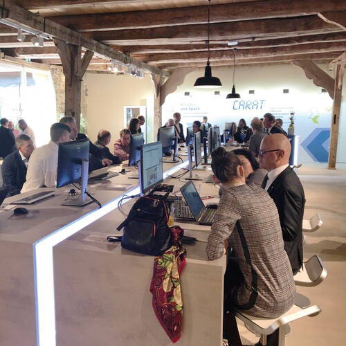 Teilnehmer eines Workshops arbeiten in einem modernen, offen gestalteten Büro gemeinsam an Computern entlang einer langen Tischreihe.