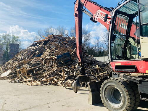 Bagger mit Greifarm sortiert einen großen Haufen ausrangierter Holzpaletten und Bretter auf einem Recyclinghof unter blauem Himmel