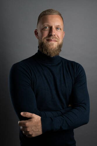 Selbstbewusster Mann mit blondem Vollbart und dunkelblauem Rollkragenpullover steht vor grauem Hintergrund und verschränkt locker die Arme.