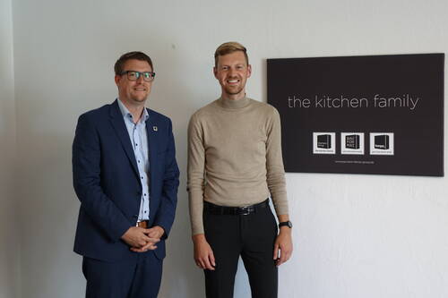 Zwei lächelnde Männer in Businesskleidung stehen in einem hellen Büro neben einem Wandposter mit der Aufschrift „the kitchen family“.