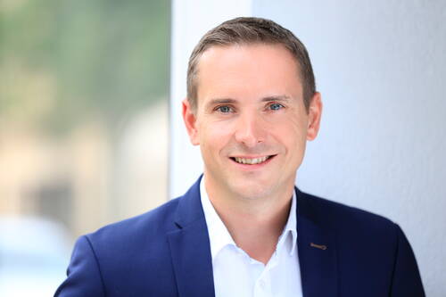 Lächelnder Mann im blauen Anzug und weißem Hemd lehnt an einer hellen Wand und blickt direkt in die Kamera