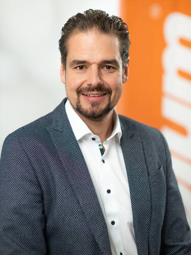 Lächelnder Mann im Business-Outfit mit dunkelblauem Sakko und weißem Hemd vor unscharfem orangefarbenem Hintergrund