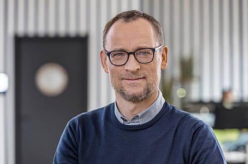 Mittelalterlicher Mann mit Brille und Bart lächelt freundlich in einem modernen Büro mit unscharfem Hintergrund